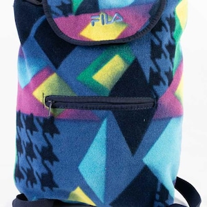 Può includere: Zaino in pile colorato con un motivo geometrico nei toni del blu, viola, giallo e verde. Lo zaino presenta una tasca frontale con cerniera, una chiusura con patta e il marchio "FILA" in azzurro. Misure non disponibili.