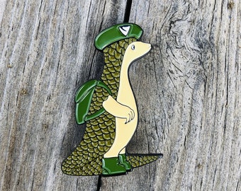 May include: A green and yellow enamel pin of a cartoon pangolin wearing a green hat with a white triangle on it. The pangolin is carrying a green backpack and has a yellow body with green scales.
