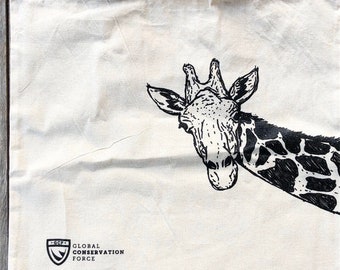May include: A white canvas tote bag with a black and white illustration of a giraffe's head peeking out from the right side. The bag has black straps and a black logo that reads "GLOBAL CONSERVATION FORCE" with a shield design.