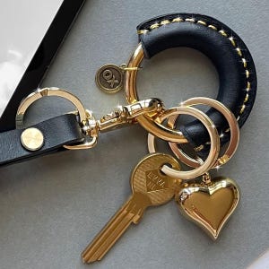 May include: A black leather keychain with gold-colored hardware. The keychain features a gold heart charm, a key, and a small round charm with "OX" on it. The leather is stitched with gold thread.