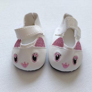 May include: A pair of white doll shoes with a cat face design. The shoes have pink glitter ears, embroidered eyes, and a pink embroidered nose and mouth. Each shoe has a white strap.