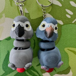 May include: Two parrot-shaped keychains. One is gray, the other is blue. Both have black beaks, yellow and black eyes, and red feet. The keychains are attached to silver key rings. The background is a green and white floral pattern.