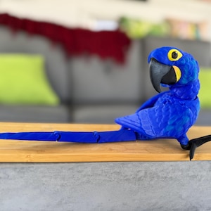 Könnte beinhalten: Eine leuchtend blaue Papageienfigur mit schwarzem Schnabel und gelben Akzenten um die Augen. Der Vogel sitzt auf einer Holzoberfläche und zeigt sein detailliertes Design und den beweglichen Schwanz.