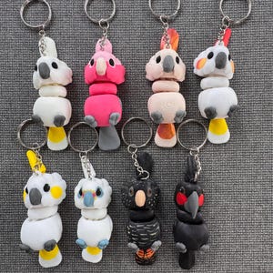 May include: A collection of handmade parrot keychains in various colors. The keychains feature detailed designs of different parrot species, including white, pink, and black parrots, each with a keyring attached. The keychains are made of clay.