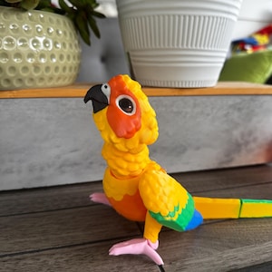 May include: A colourful parrot figurine with a yellow body, orange chest, and green and blue wing details. The bird has a black beak, a white eye with a brown pupil, and pink feet. The figurine is displayed on a wooden surface.