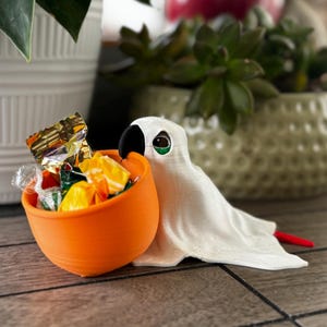 Könnte beinhalten: Ein weißer, geisterförmiger Süßigkeitenhalter mit einer orangefarbenen Schüssel voller verpackter Süßigkeiten. Der Geist hat einen schwarzen Schnabel, ein grünes Auge und einen roten Schwanz. Die Schüssel ist mit verschiedenen bunten Süßigkeiten gefüllt.