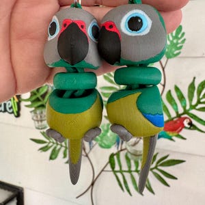 May include: Two parrot-shaped keyrings. Each keyring features a grey head with red accents, a black beak, and blue eyes. The body is yellow and green, with a grey tail. The keyrings are connected to a silver chain.
