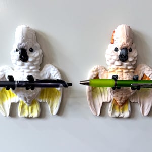 May include: Two white cockatoo-shaped pen holders with yellow and orange accents. One holds a black pen, the other a green pen. The birds have black beaks and eyes, and are mounted on a white surface.