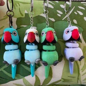 May include: Four colorful parrot keychains. Each keychain features a parrot with a red beak, black accents, and a body in shades of blue, green, white, and purple. The keychains are hanging against a green leaf pattern.