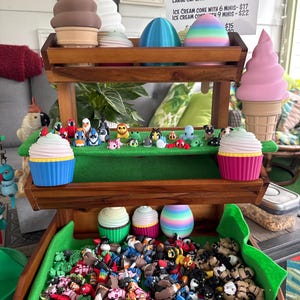 May include: A wooden display stand filled with colorful 3D-printed fidget toys. The stand features cupcake and ice cream cone-shaped containers, each holding various mini fidgets. The sign reads "MINI 3D PRINTED FIDGETS IN CONTAINER OF YOUR CHOICE!"