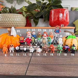 Puede incluir: Una colección de veintisiete pequeñas figuras coloridas con temática de Halloween dispuestas sobre una superficie. Las figuras incluyen varios personajes como fantasmas, esqueletos, monstruos y animales, cada uno marcado con un número del uno al veintisiete.