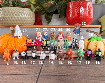 Puede incluir: Una colección de veintisiete pequeñas figuras coloridas con temática de Halloween dispuestas sobre una superficie. Las figuras incluyen varios personajes como fantasmas, esqueletos, monstruos y animales, cada uno marcado con un número del uno al veintisiete.