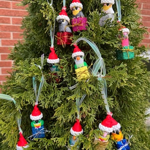 May include: A collection of handmade Christmas ornaments shaped like parrots, each wearing a red Santa hat. The parrots are perched on miniature gift boxes wrapped in various colors. The ornaments are suspended from a green evergreen tree.