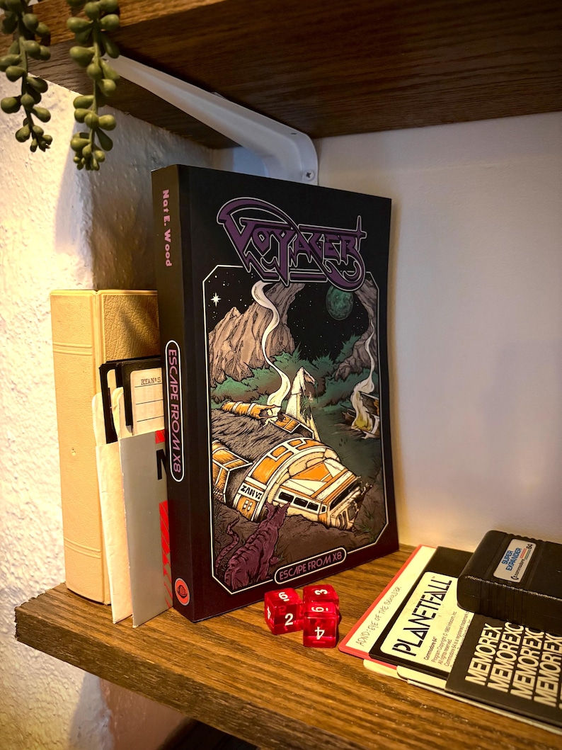 May include: A black book titled Voyager: Escape from X8 by Nat E. Wood with a graphic illustration of a crashed spacecraft on a dark landscape. The book is positioned next to a stack of floppy disks and two translucent red six-sided dice.
