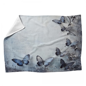May include: A soft, light blue throw blanket featuring a pattern of blue butterflies. The butterflies are in various sizes and positions, with detailed wing patterns. The blanket has a textured, distressed background, and a folded corner.