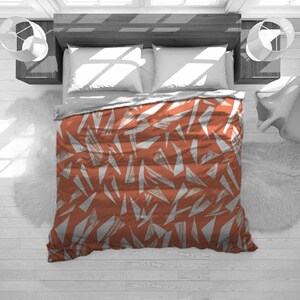 May include: A bed with a rust-colored duvet cover featuring a white geometric pattern of irregular shapes. The bed has white pillows and a gray top sheet. The room has a modern design with white walls and a wooden bed frame.