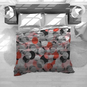 May include: A bed with a gray comforter featuring a pattern of overlapping circles in shades of gray, black, and red. The bed has white pillows and is flanked by wooden nightstands with lamps. The room has a modern, minimalist aesthetic.