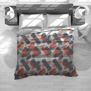May include: A bed with a gray comforter featuring a repeating pattern of gray and coral-colored abstract shapes. The bed is made with white pillows and a gray bed skirt. The room has a modern design with white walls and wooden furniture.