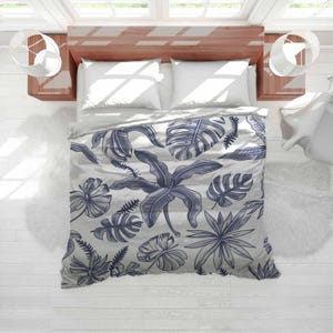 May include: A bed with a white duvet cover and pillows, showcasing a grey and blue botanical pattern of leaves and flowers. The bed is set against a light wood headboard and white walls.