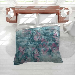 May include: A teal and pink floral duvet cover on a bed with white pillows. The bedding has a watercolor-like design with abstract pink flowers and a textured, distressed appearance. The bed is set against a wooden headboard and white walls.