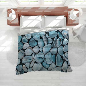 May include: A duvet cover featuring a pattern of blue and gray stones. The stones vary in size and shape, creating a natural, textured appearance. The bedding is set on a wooden bed frame with white pillows and a white sheet.