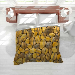 May include: A bed with a duvet cover featuring a pattern of yellow, brown, and gray stones. The stones are arranged closely together, creating a textured appearance. The bed has white pillows and is set against a light wood headboard.