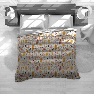 May include: A bed with a white comforter featuring a watercolor pattern of autumn leaves in shades of yellow, orange, and red. The bed is made up with white pillows and a gray sheet. The bed is on a white wooden floor.