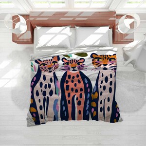 May include: A colorful duvet cover featuring three stylized cheetahs. The cheetahs have spots and are in shades of pink, orange, and navy blue. The background is white with some green and pink leaf shapes.