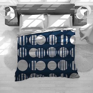 May include: A navy blue and silver patterned duvet cover on a bed. The design features large silver circles, some with vertical stripes, against a dark blue background. White pillows and a light gray top sheet are visible.