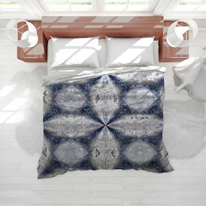 May include: A duvet cover with a symmetrical pattern in grey and blue, with floral and geometric shapes. The bedding is on a bed with white pillows and a wooden headboard. The overall aesthetic is elegant and decorative, suitable for a bedroom.
