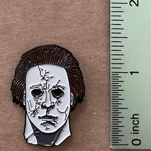 May include: Enamel pin featuring the mask of Michael Myers from the Halloween horror film series. The pin is white with black details and brown hair. The pin measures approximately 1 inch in height.