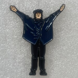 May include: Enamel pin depicting a figure in a navy blue coat with arms outstretched. The figure is wearing a black hat and black pants. The pin has gold accents on the hands and face.