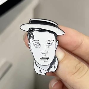 Puede incluir: Pin de esmalte blanco y negro de un hombre con un sombrero blanco. El hombre tiene el pelo corto y oscuro y una expresión seria.