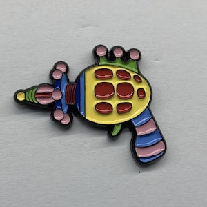 May include: A brightly coloured ray gun pin with a yellow body, red accents, and a black outline. The pin features pink, blue, green, and yellow details, and is likely made of enamel or a similar material.