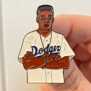 May include: A black and white enamel pin of a baseball player wearing a white jersey with the word "Dodgers" in blue lettering. The player has a black headband and is wearing a gold chain.