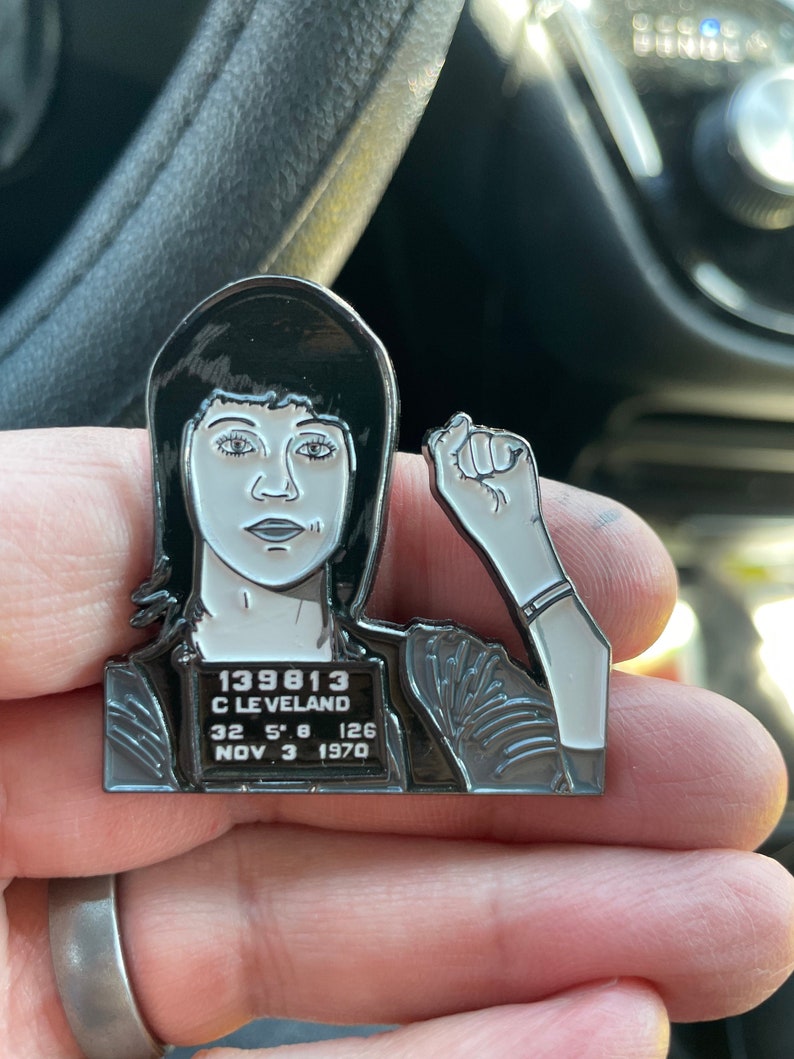 May include: Black and white enamel pin of a woman with short black hair. The pin features a mugshot style image with the text "139813 C LEVELAND 32 5'8 126 NOV 3 1970".