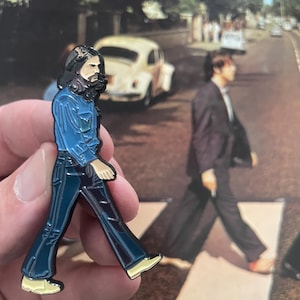 Puede incluir: Pin esmaltado que representa a un hombre con camisa azul y pantalón azul oscuro, caminando. El pin es una representación estilizada de una figura de la icónica portada del álbum Abbey Road. El fondo muestra un coche amarillo y otra figura caminando.
