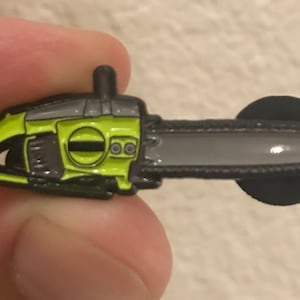 Puede incluir: Un pin de esmalte verde y negro con forma de motosierra. La motosierra tiene un mango negro y una hoja plateada.