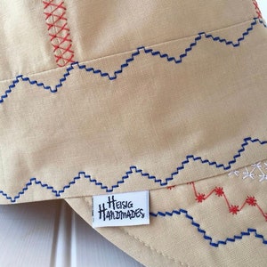 May include: A beige fabric hat with blue and red stitching. The hat has a label that says "Heisig Handmades".