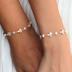 May include: Two gold bracelets with delicate white pearl and crystal leaf accents. The bracelets are worn on the wrists of two people holding hands.