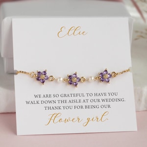 Könnte beinhalten: Ein goldfarbenes Armband mit drei lila blütenförmigen Edelsteinen und kleinen Perlen. Das Armband liegt auf einer weißen Karte mit dem Namen "Ellie" und dem Text "Flower girl."