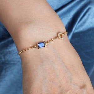 Könnte beinhalten: Ein goldenes Kettenarmband mit einem blauen Edelstein und einem kleinen, runden Anhänger mit klaren Steinen.