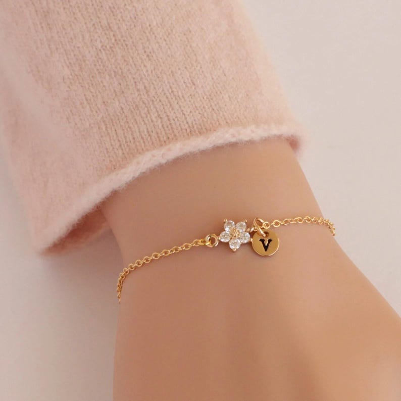 May include: A gold chain bracelet with a small flower charm and a round charm with the letter "V" on it.