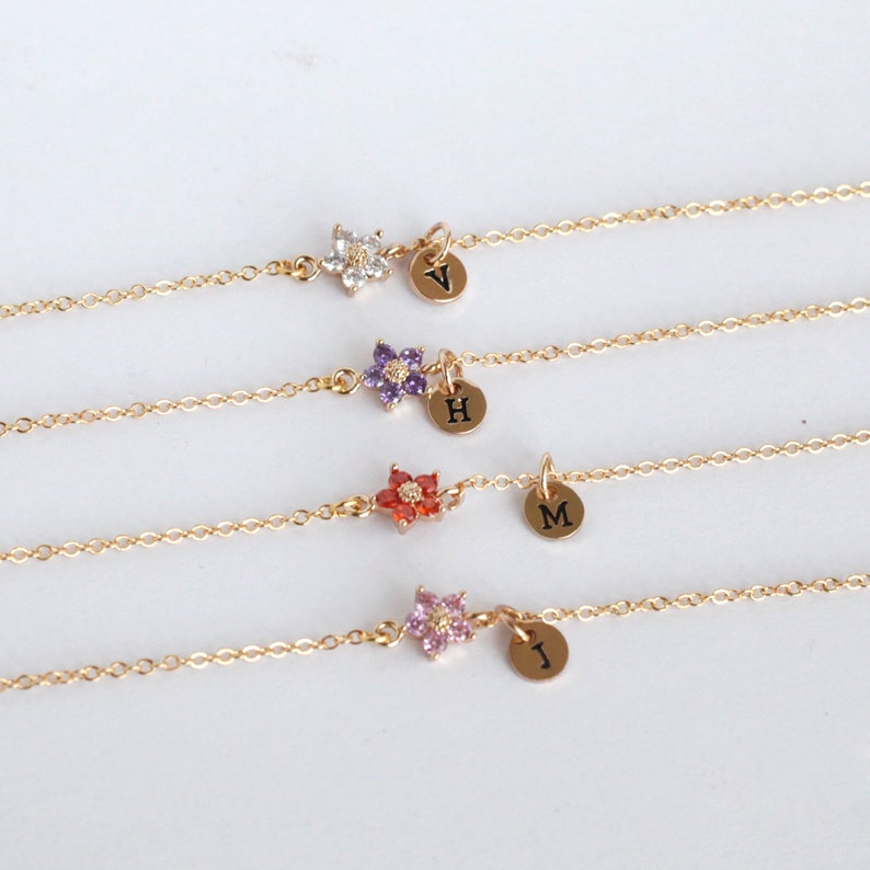May include: Four gold chain bracelets with a small flower charm and a personalized initial charm. The flower charms are in different colors: white, purple, red, and pink.