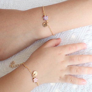 May include: Two gold bracelets with charms. The bracelet on the left has a purple flower charm and a gold disc with a letter 'H' on it. The bracelet on the right has a pink flower charm and a gold disc with a letter 'J' on it.