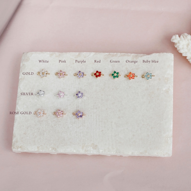 May include: A display of flower charm connectors in various colors, including white, pink, purple, red, green, orange, and baby blue. The charms are available in gold, silver, and rose gold finishes.