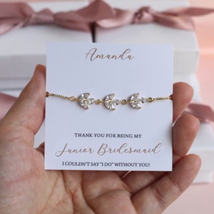 May include: A white card with the text "Amanda" at the top, followed by "THANK YOU FOR BEING MY Junior Bridesmaid I COULDN'T SAY 'I DO' WITHOUT YOU!"  A gold bracelet with three crystal leaf charms is displayed on the card.