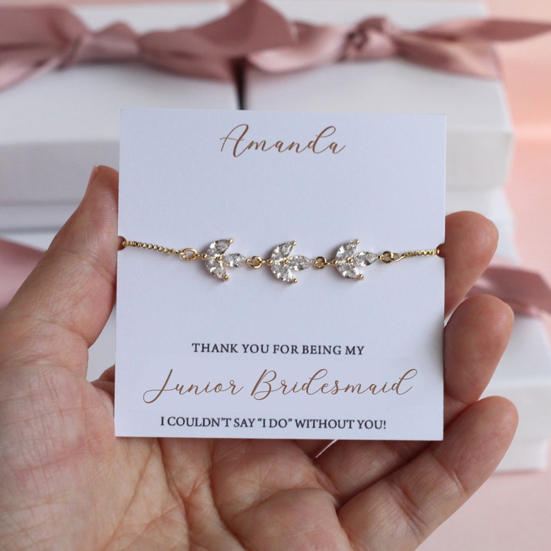 May include: A white card with the text "Amanda" at the top, followed by "THANK YOU FOR BEING MY Junior Bridesmaid I COULDN'T SAY 'I DO' WITHOUT YOU!"  A gold bracelet with three crystal leaf charms is displayed on the card.