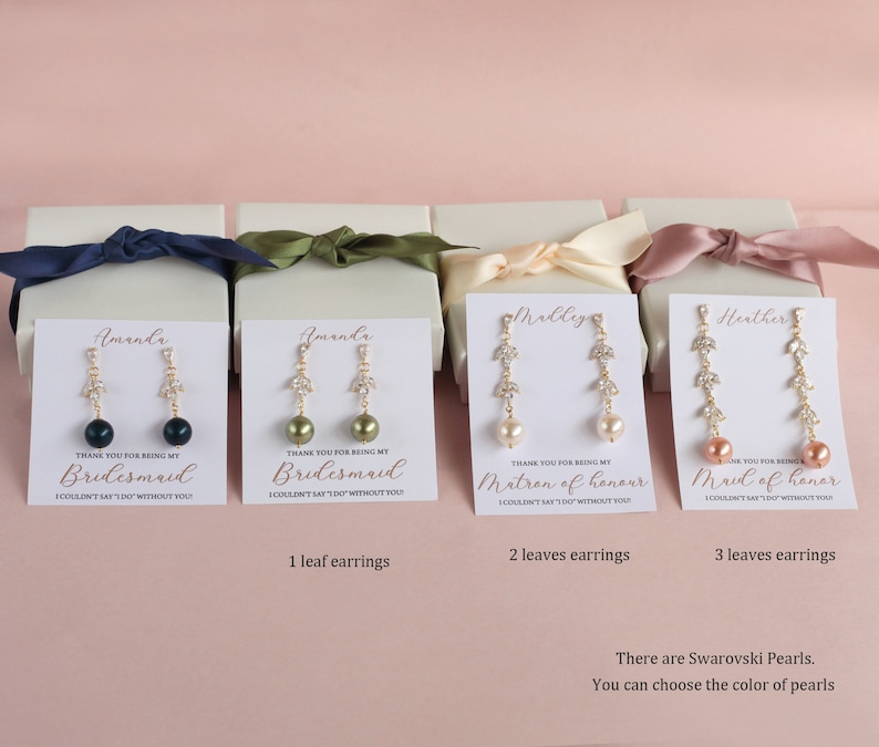 May include: Four white gift boxes with gold lettering and a pair of gold earrings with a pearl and crystal leaves in each box. The boxes are labeled "Bridesmaid", "Bridesmaid", "Matron of Honor", and "Maid of Honor". The text "There are Swarovski Pearls. You can choose the color of pearls" is at the bottom of the image.