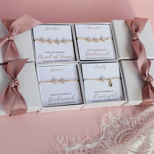 May include: Four white gift boxes with pink ribbon bows, each containing a gold bracelet with a message card. The cards read "Thank you for being my Maid of Honor", "Thank you for being my Bridesmaid", "Thank you for being my Bridesmaid", and "Thank you for being my Flower Girl".